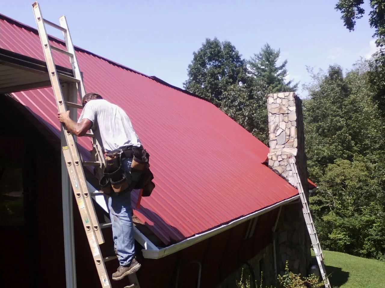 Roofer on a ladder beside a red metal roof and stone chimney — Soffit & Fascia Repair by Roofing Brothers in Central Falls